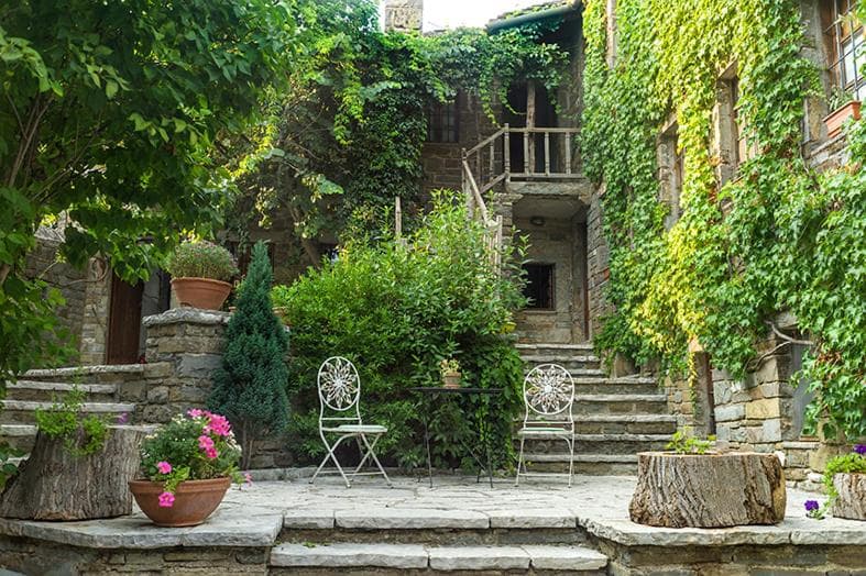 Stone courtyard with ivy-covered houses, potted plants, and white metal garden furniture.