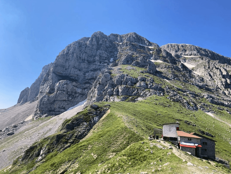 Stone mountain hut on grassy slope beneath massive rocky peaks under clear blue sky