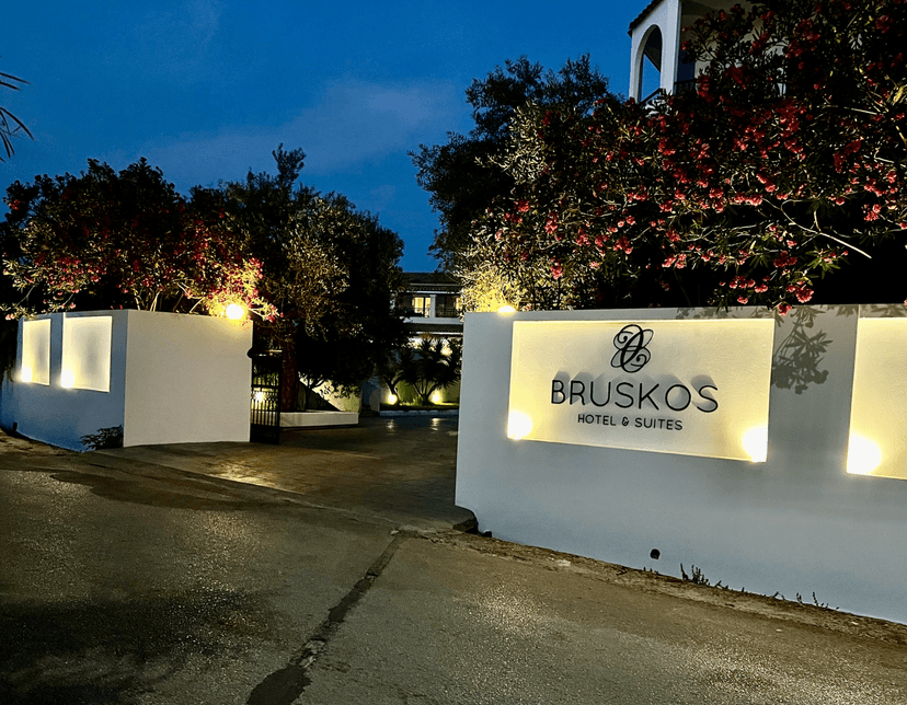 Entrance to Bruskos Hotel & Suites at night with illuminated sign and flowering trees.