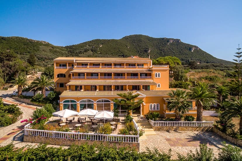 Orange hotel building with outdoor dining patio, palm trees, and green mountains under blue sky.