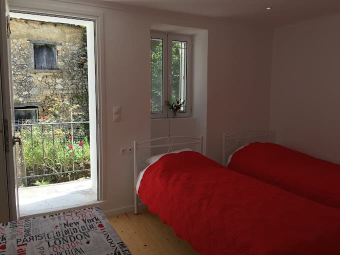 Twin beds with red covers in a white room opening to a stone building and garden.