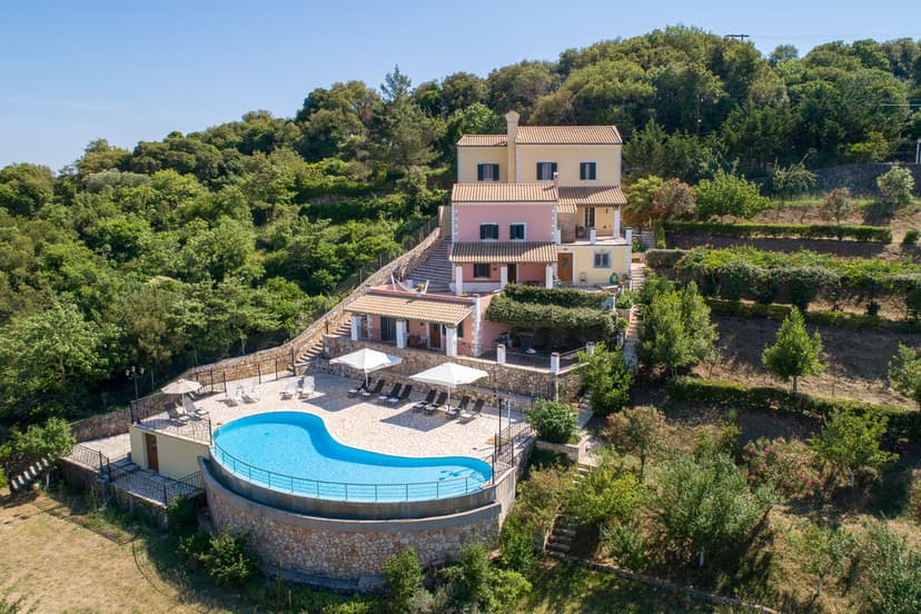 Multi-level villa with curved pool on terraced hillside near dense green forest in Corfu.