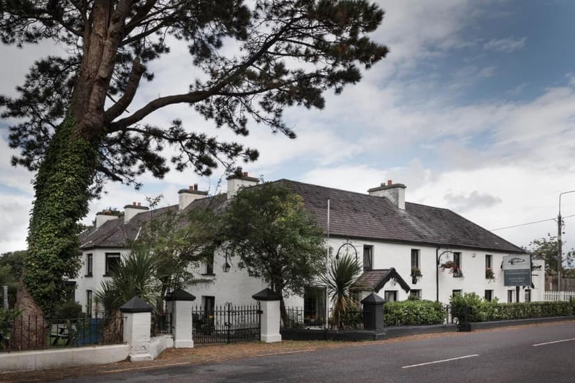 White, whitewashed hotel building with dark roof, large tree, and iron fence beside a road.