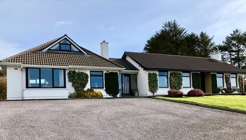 Derrynane Bay House, a white bungalow with dark trim and ivy, faces a gravel driveway.