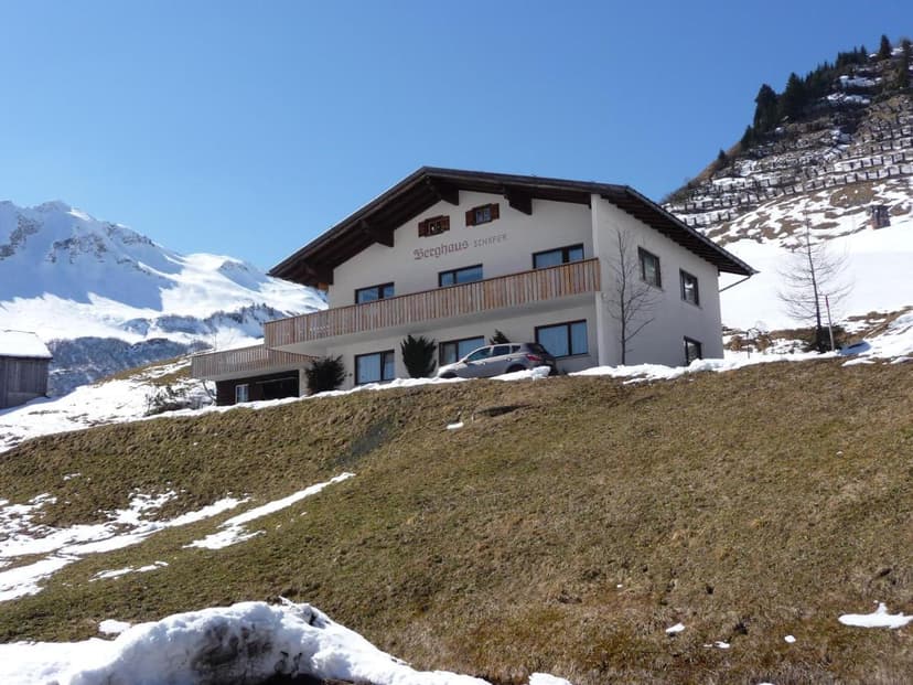 Berggasthaus Schäfler building on grassy slope with melting snow and snowy mountains.
