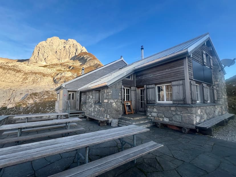 Zwinglipasshütte mountain hut with wooden benches on stone patio, massive rock peak in background.
