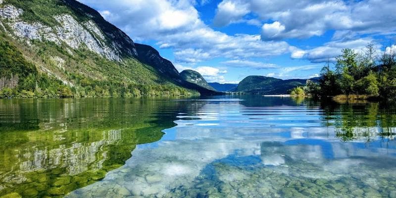 Clear water of Lake Bohinj reflecting mountains and blue sky with white clouds