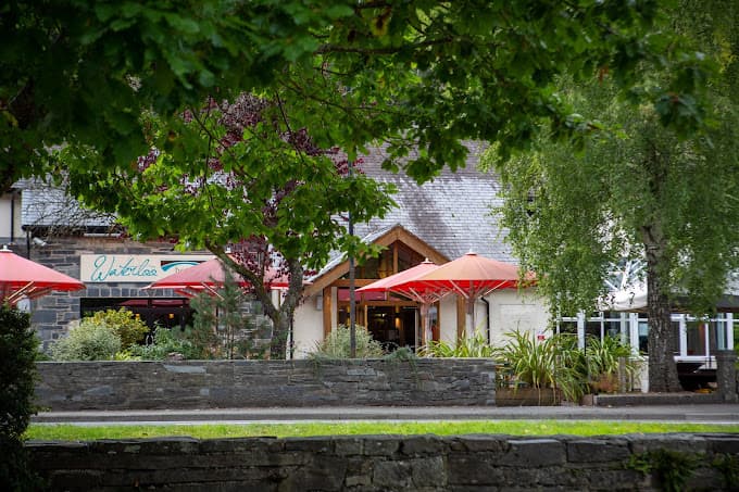 The Waterloo Hotel entrance with red umbrellas, seen through lush green foliage and over a stone wall.