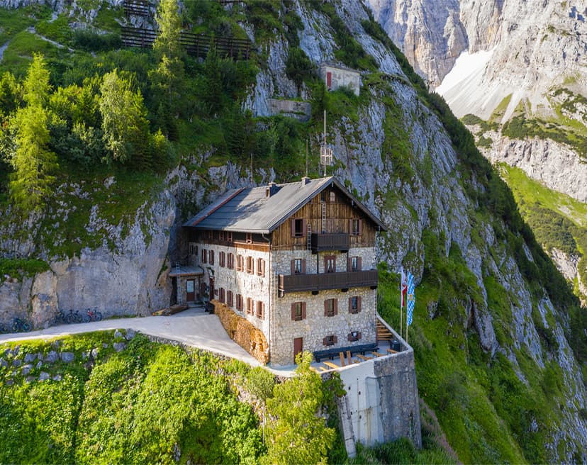 Stone mountain hut built into a steep, green mountainside with rocky peaks above
