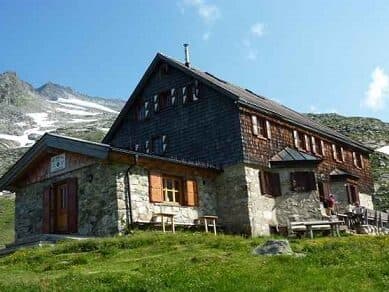 Stone and wood mountain hut on grassy slope with snow-capped peaks under blue sky