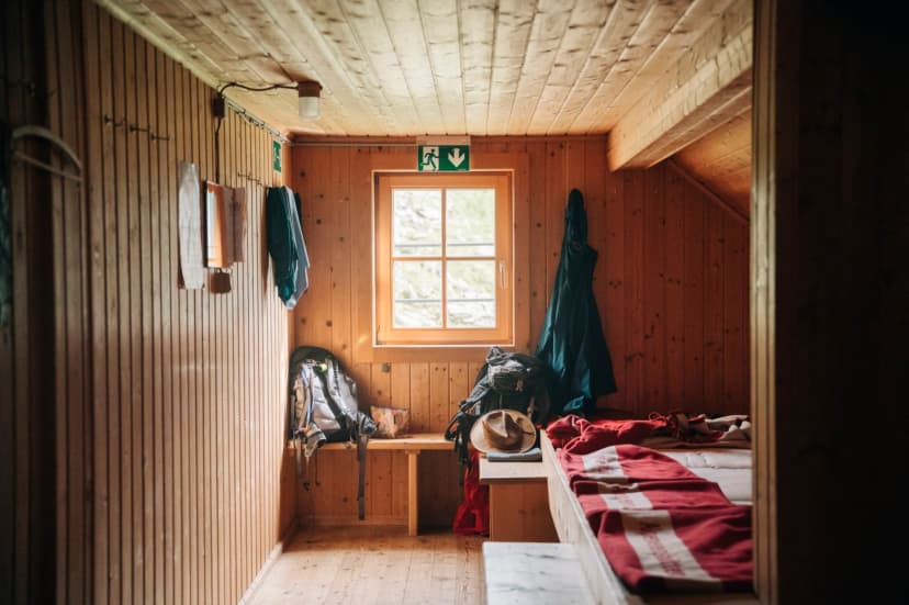 Interior of wooden mountain hut with bunks, backpacks, and window showing greenery