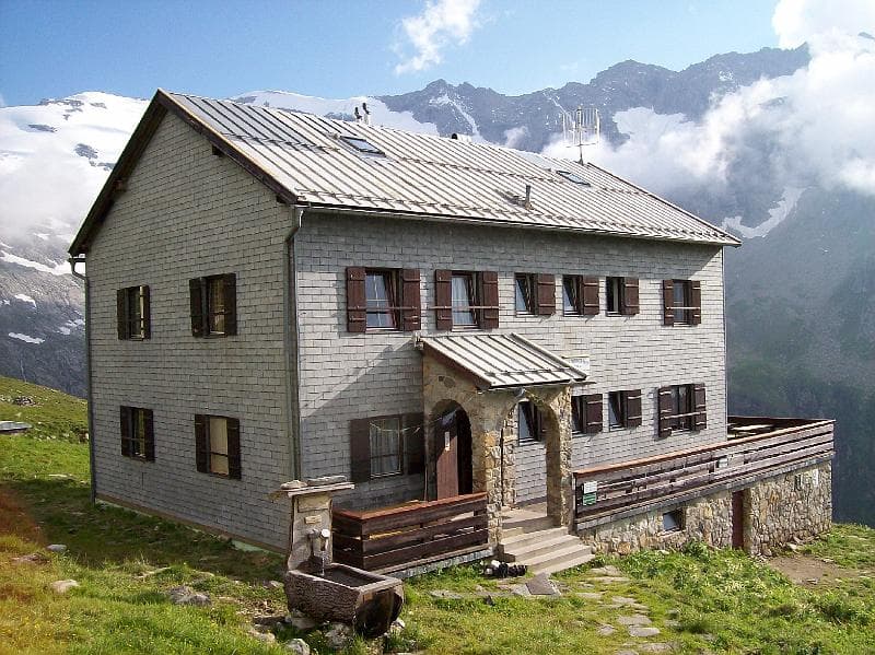 Mountain hut with stone base and slate siding against snow-capped peaks and clouds