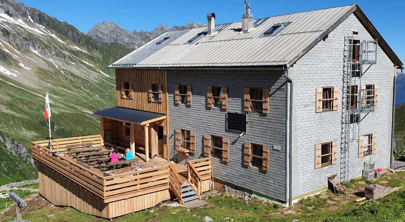 Mountain hut with wooden deck and outdoor seating against steep green slopes