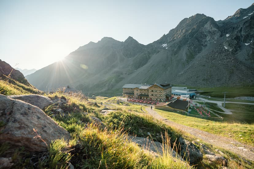 Mountain hut in alpine valley with sun flare over rocky peaks and green meadow.