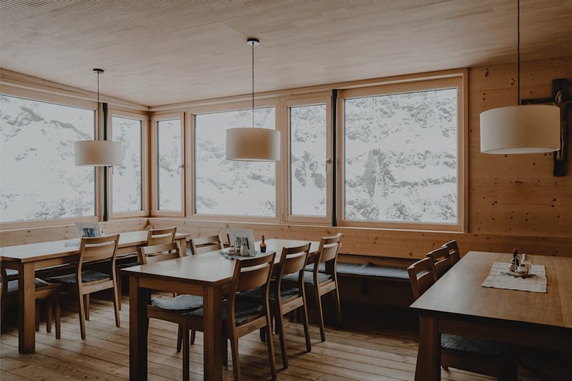 Dining room with wooden tables and chairs overlooking snowy mountains through large windows at Winnebachseehütte.