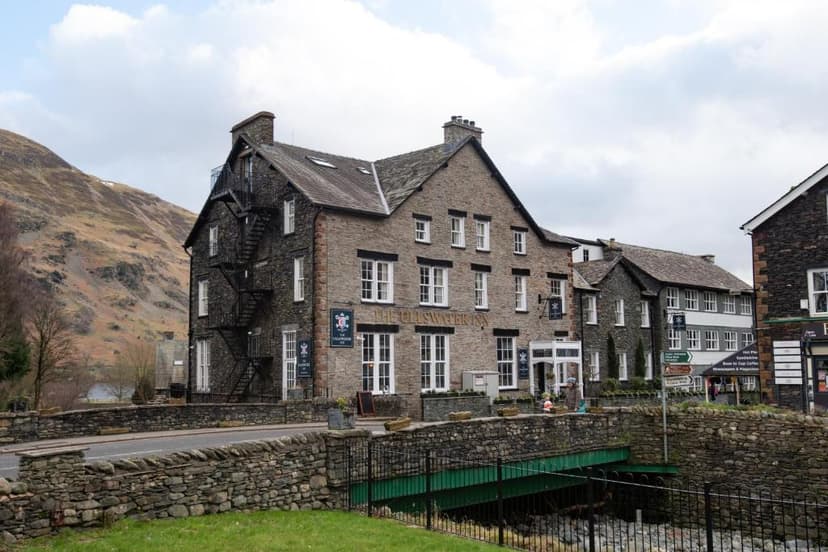 The Ullswater Inn stone building next to a bridge with a grassy bank and brown mountain backdrop.
