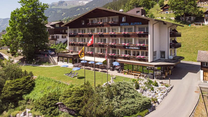 Hotel Alpina restaurant in Grindelwald with Swiss flags and green lawn against mountain backdrop.