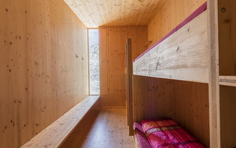 Interior of a wooden mountain hut room with bunk beds and view of rocky terrain through a narrow window.