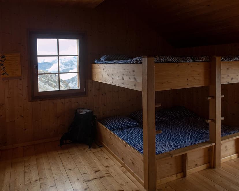 Wooden bunk beds inside a mountain hut with a view of rocky peaks and clouds.