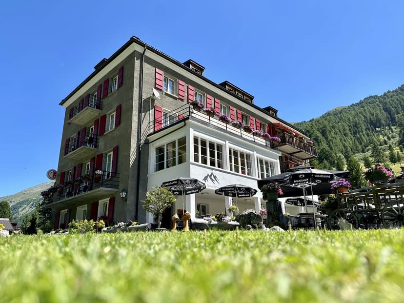 Hotel Schwarzhorn with red shutters, outdoor seating, and green mountain backdrop under blue sky.