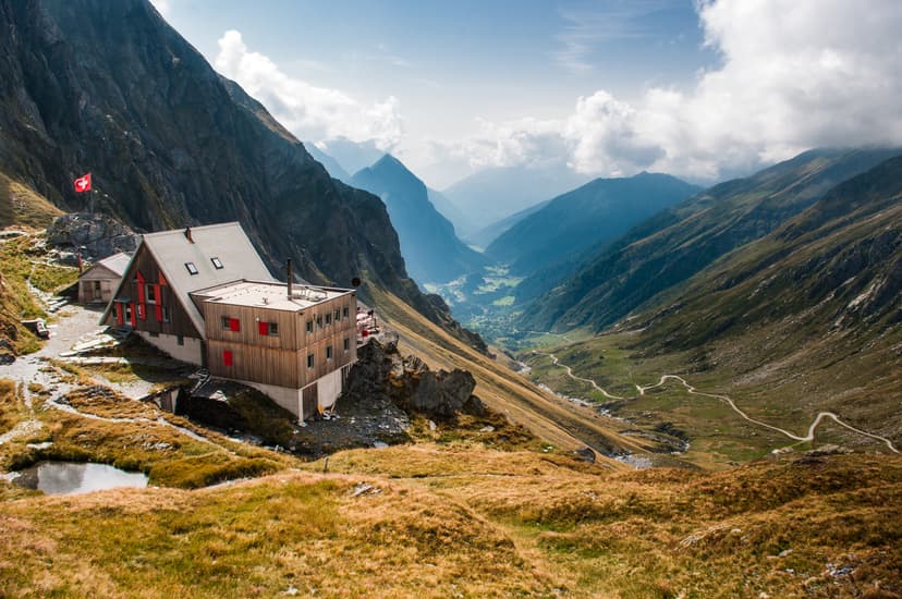 Mountain refuge hut with Swiss flag overlooking deep valley and winding road in the Alps