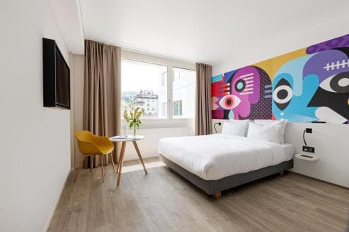Modern hotel room with colorful mural, white bedding, and view of St. Gallen buildings.