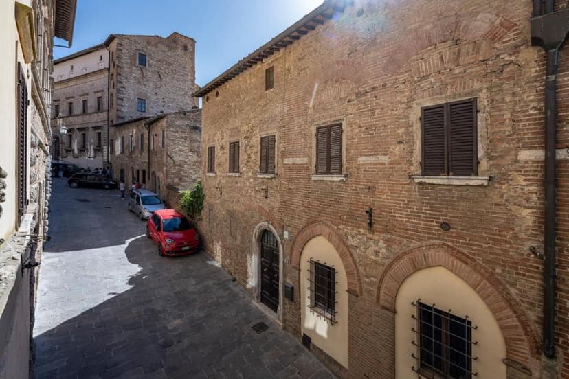 Narrow cobblestone street with parked cars alongside old brick and stone buildings under bright sun.