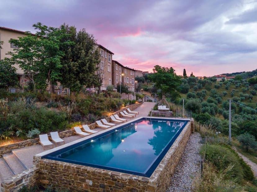 Swimming pool with lounge chairs by stone wall overlooking olive groves at sunset.