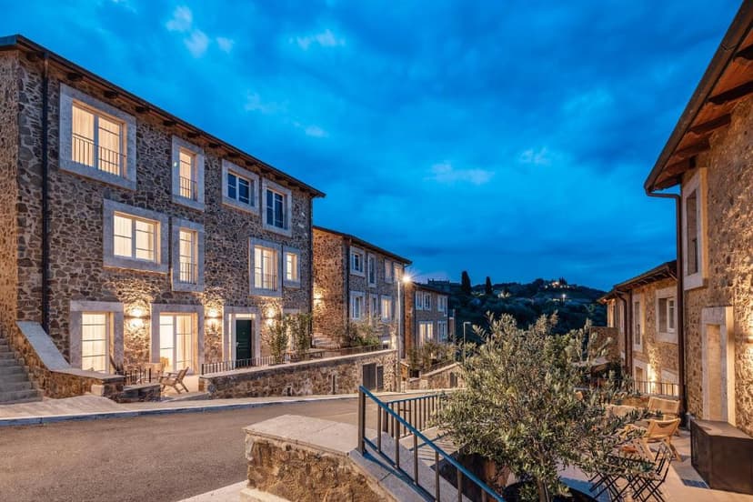 Stone buildings illuminated at dusk along a street, with hills in the background, Corte Francigena.