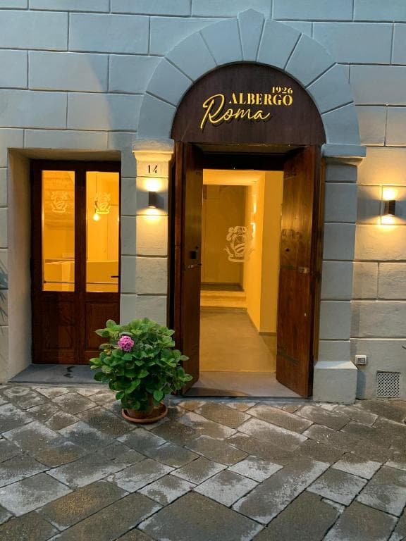 Entrance to Albergo Roma with illuminated sign, wooden doors, and potted plant on stone pavement.