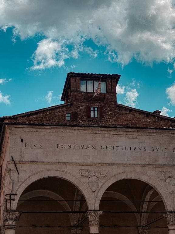 Stone arcade with Latin inscription under a brick tower against a bright blue cloudy sky.