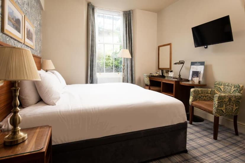 Hotel bedroom with white bedding, patterned armchair, desk, and window view.