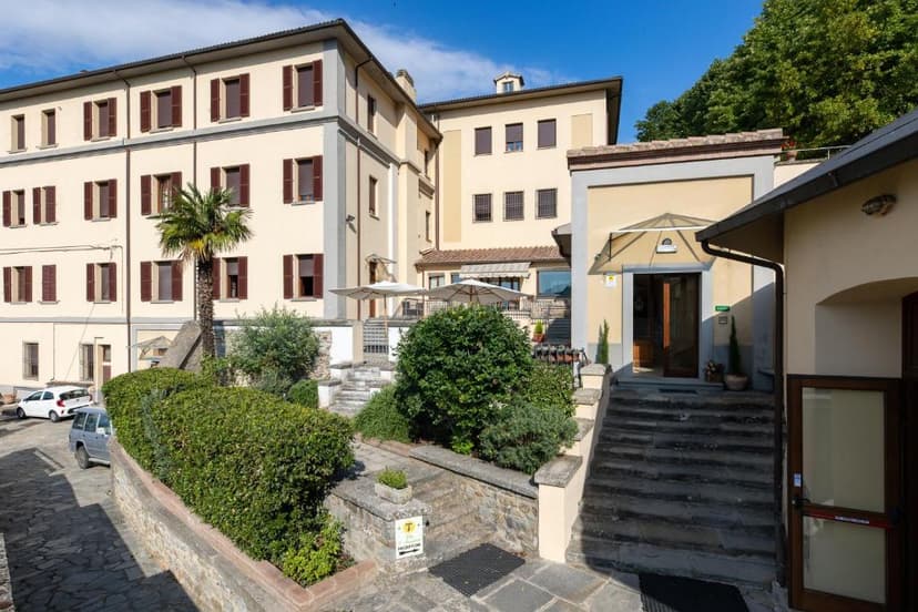 Palazzo del Mercante building entrance with stone steps, lush greenery, and parked cars on a sunny day.