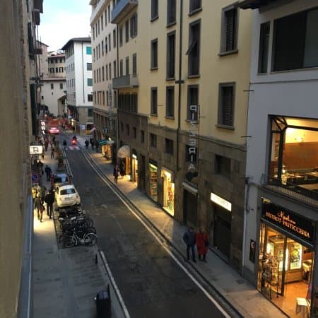 Narrow street in Florence with historic buildings, shops, and people walking at dusk.