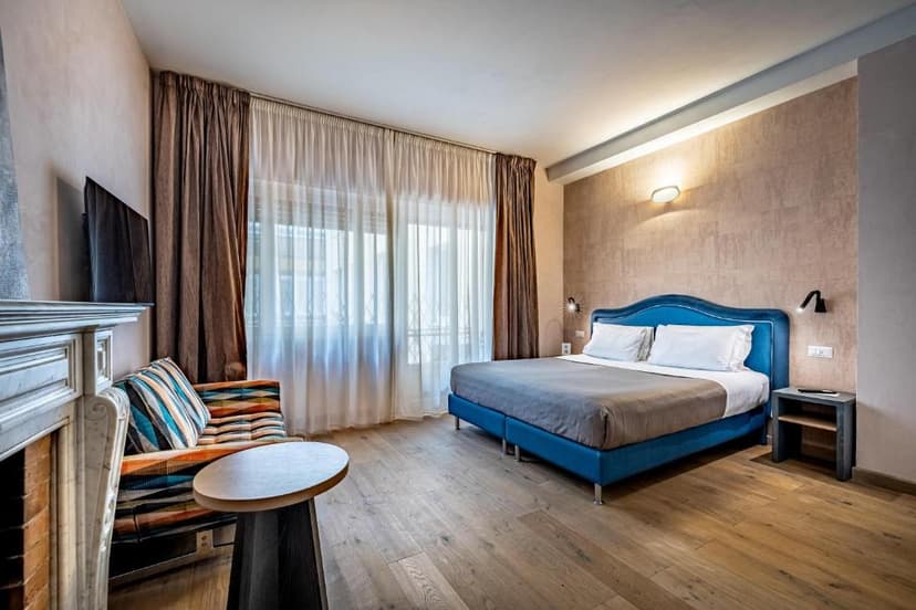 Bedroom with blue velvet bed, fireplace, striped chair, and large window with sheer curtains.
