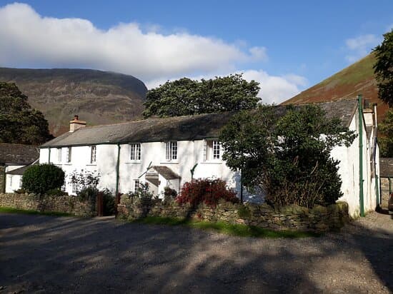 White farmhouse with stone wall, set against large mountains under blue sky.