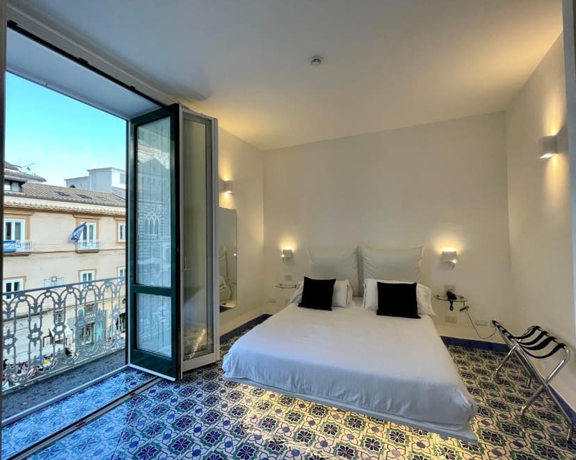 Bedroom with white bedding, blue patterned tile floor, and balcony view of Duomo.