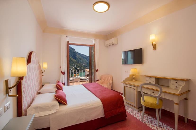 Hotel room with bed, desk, and balcony view of steep green mountains and water.
