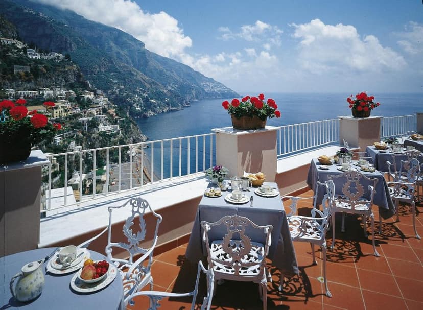 Outdoor dining terrace with white metal chairs overlooking steep coastal mountains and the sea.