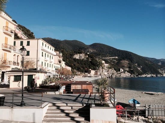 Seaside promenade with buildings, beach, and steep green hills under a clear blue sky at Locanda A Cà Du Gigante.