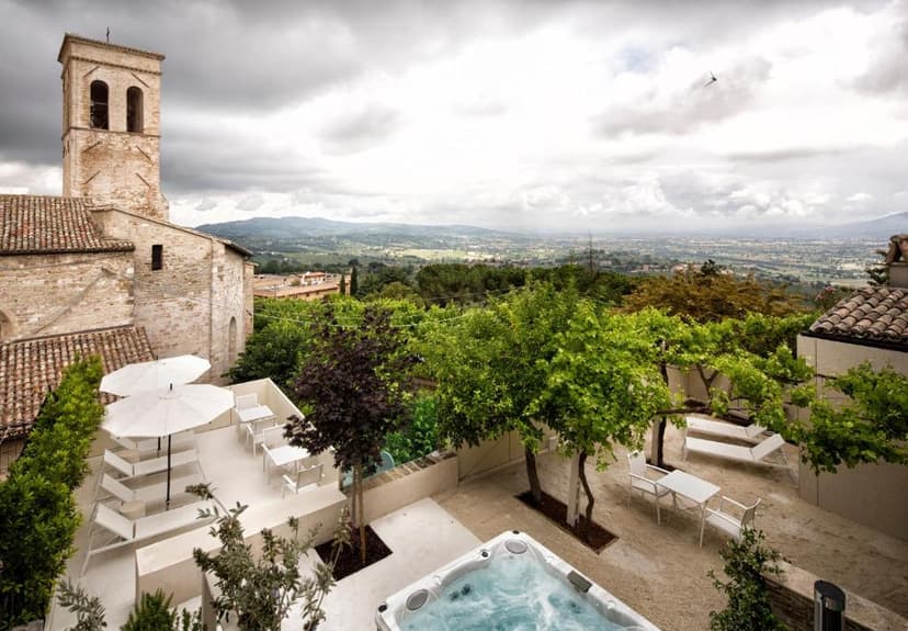Terrace with hot tub, white furniture, and bell tower overlooking green valley under cloudy sky.