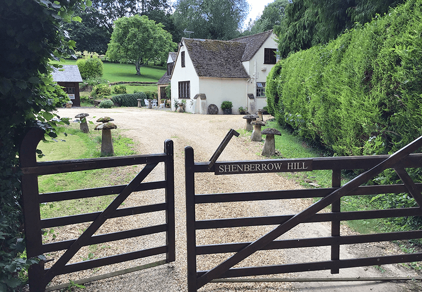 Open wooden gate marked Shenberrow Hill leading to a cream cottage and gravel drive.