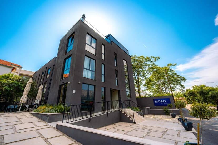 Modern dark gray building exterior with large windows and a blue "MORO INN KORČULA" sign under a bright blue sky.