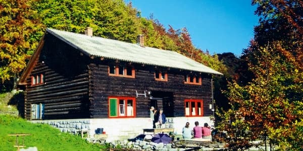 Dark log mountain hut with people sitting outside against an autumn forest backdrop