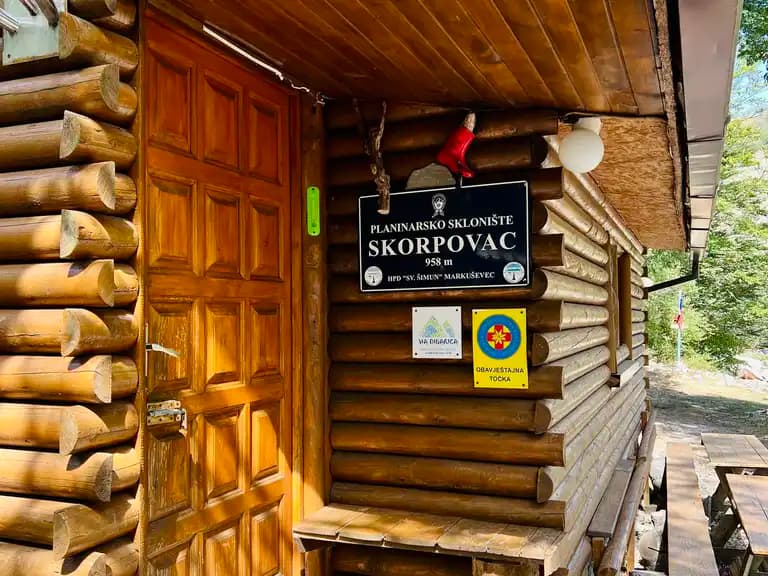 Log cabin entrance with "Skorpovac" sign, wooden door, and outdoor seating in a wooded area.