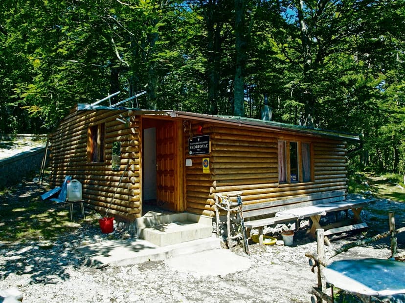 Log cabin shelter with "Skorpovac" sign surrounded by dense green forest on a sunny day.