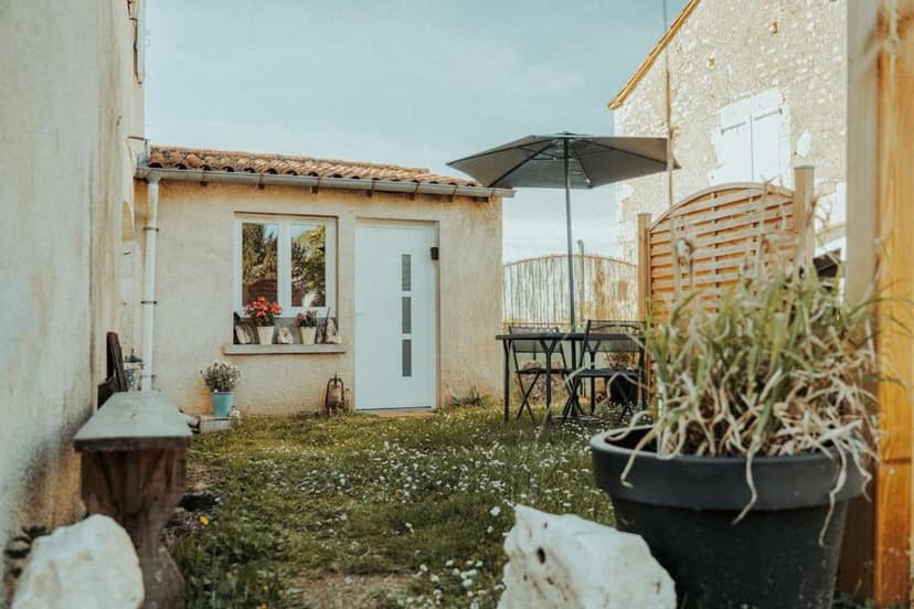 Outdoor patio area with small building, grass, potted plants, and outdoor dining set under an umbrella.