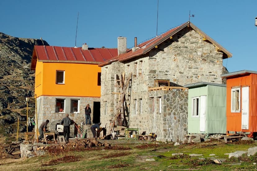 Mountain hut complex with stone and bright orange buildings, people working outdoors near logs.
