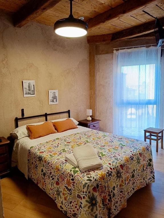 Bedroom with floral bedspread, exposed wooden beams, and sheer curtains in Sahagún.