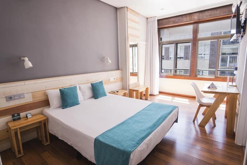 Hotel room with white bedding, teal accents, wood furniture, and city view windows in Hotel Alda Centro Ponferrada.